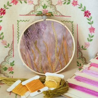 Embroidery hoop contains grass stitched onto a purple photographic background, surrounded by various threads and embroidery tools.