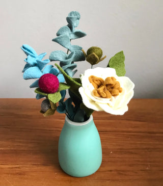 Bouquet of a variety of flowers and leaves made from felt, arranged in a small blue vase.