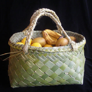 A shopping basket or bag made from woven green NZ Flax (harakeke), filled with fruit against a black backdrop.