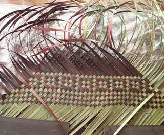 Progress shot of kete whakairo, with green and red flax woven together in tight grid pattern.
