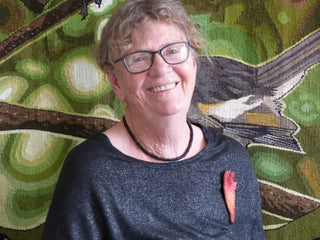 Artist and tutor of Tapestry Weaving, Marilyn Rea-Menzies smiles in front of her tapestry artwork of a New Zealand Pīwakawaka (fantail).