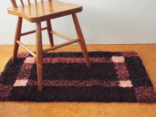 An Heirloom Hooked Fleece Rug made with red and brown wool spread out on the floor underneath a wooden chair.