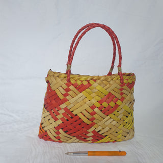 Traditional Māori Kete basket woven with light brown, red, orange and yellow Harakeke (NZ Flax), next to a pen for scale.