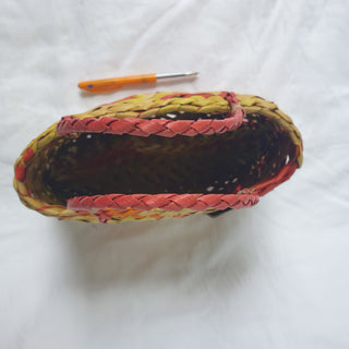 Top-view of a traditional Māori Kete basket woven with light brown, red, orange and yellow Harakeke (NZ Flax), next to a pen for scale.