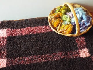 A basket of dyed wool sits atop a red and dark brown Heirloom Hooked Fleece Rug laid out on the floor.