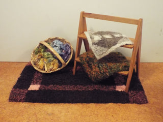 An Heirloom Hooked Fleece Rug made with red and brown wool spread out on the floor underneath a basket of dyed wool bundles and small step ladder. Two small patterned rugs have been placed on the steps.